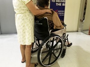 An elderly woman sits on a wheelchair, while her companion assists behind. Taken inside a hospital on March 2017.