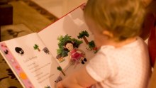 A toddler dressed in pyjamas sits on the floor pointing to a picture of a sheep on the page of a nursery rhyme book.