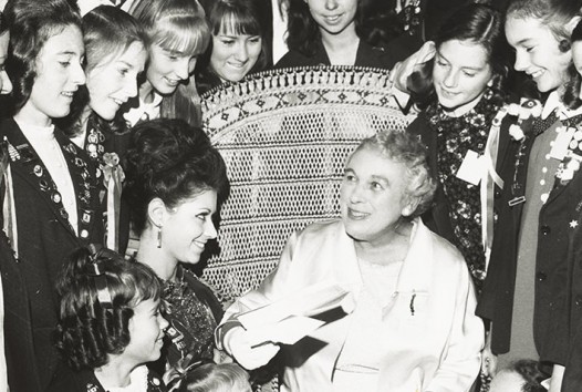 Young women surround a chair in which a beaming older woman sits as she is handed a book