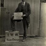 Man with hat holding De Vrije Socialist, standing with one foot on IWW San Diego