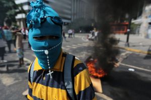 A youth wearing a mask and rosary stands near a burning barricade  in Caracas.