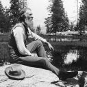 John Muir, full-length portrait, facing right, seated on rock with lake and trees in background, circa 1902. Photo: unknbown via Library of Congress (Public Domain).