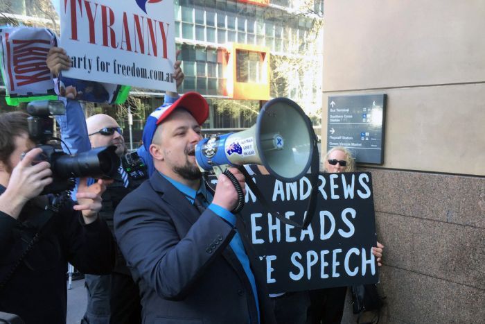 The UPF's Neil Erikson yells into a megaphone while holding a "Dan Andrews beheads free speech" sign outside court.