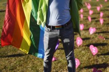 A man stands holding a rainbow flag in front of pink paper hearts decorating a grassy space.