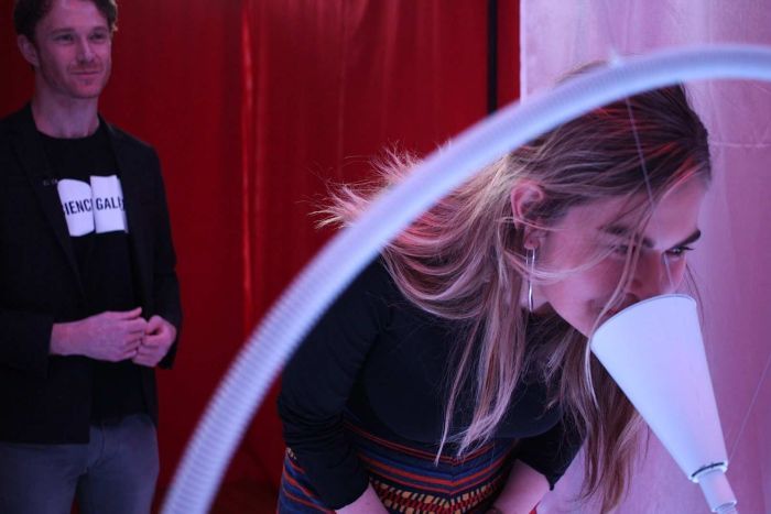 The Mix reporter Lisa Skerrett leans down and sniffs a while cone-shaped object as part of an exhibition.