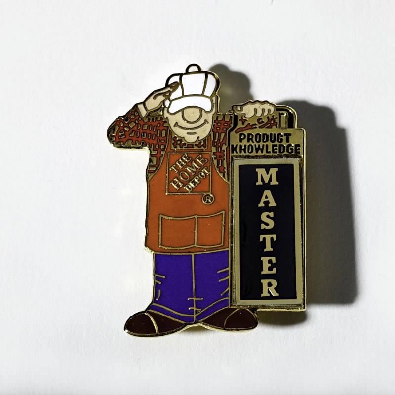 An enamel pin with a cartoon of a Home Depot employee holding a sign that reads “Product Knowledge Master”