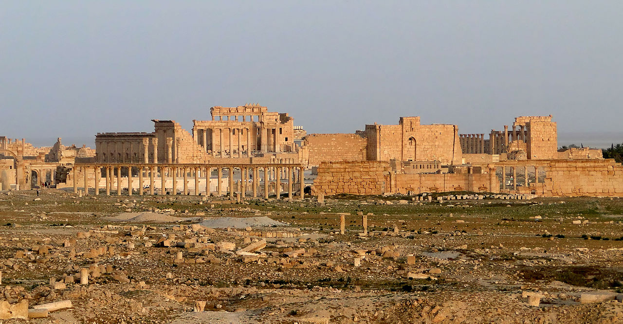 A landscape depicting ancient ruins. This is the Temple of Bel, in Palmyra. In the foreground, a field of stray stones and grass. A large structure of columns and walls made tan colored stone appears in the distance, glowing in the setting sun.