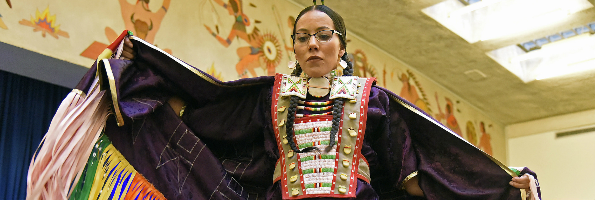 A Native American woman in elaborate traditional clothes dances in front of a Native American mural in a large room.