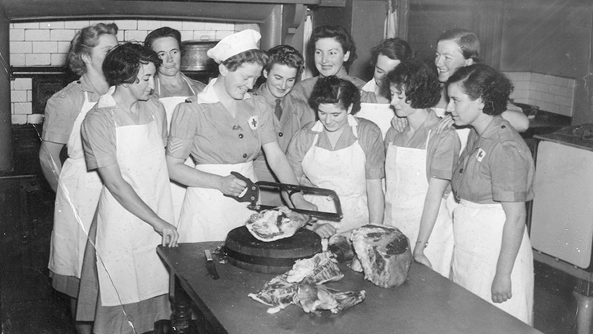 Uniformed women in aprons stand around a table while one saws a large piece of meat