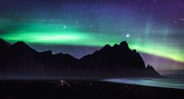 A pair of Japanese travellers watch from a black sand dune as the northern lights dance over Vestrahorn Mountain in East ...