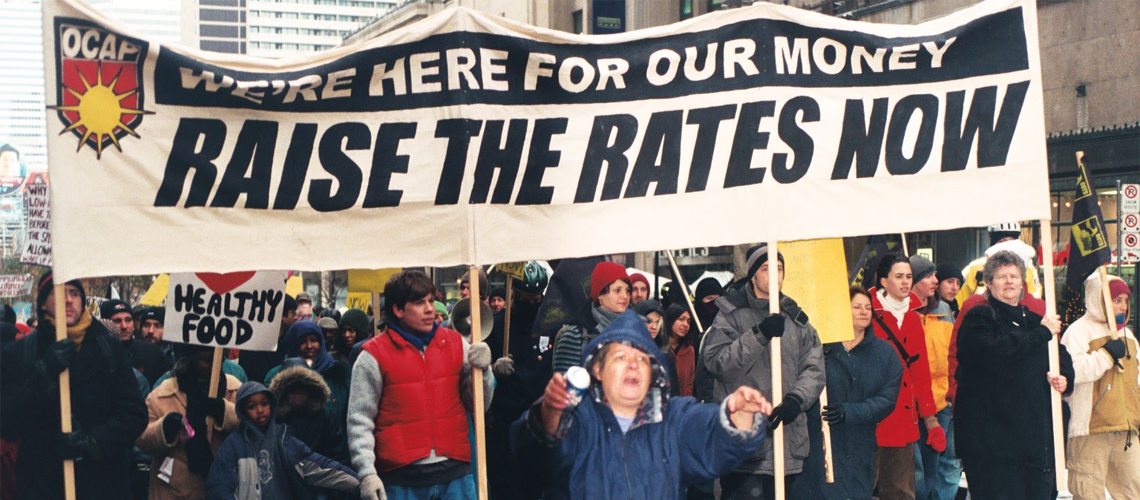 Raise the Rates march. Large crowd following behind a banner that says "We're here for our money, Raise the Rates Now".