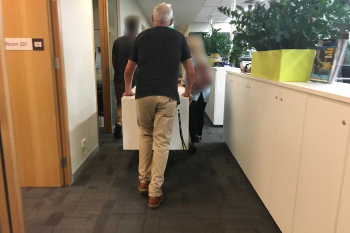 A man whose back is towards the camera walks through an office. Two other people in are blocked out.