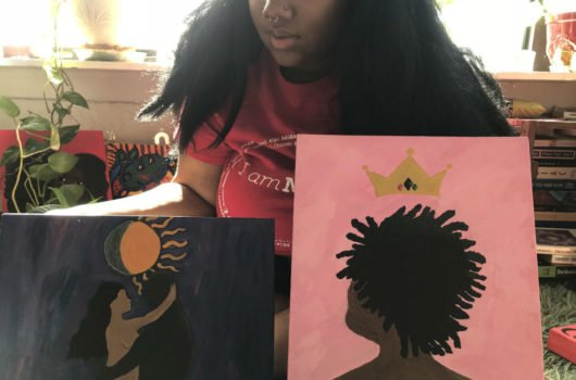 [Photo: A Black woman sits holding two vibrant paintings, one dark blue, one pink. Only the bottom half of her face is visible.]