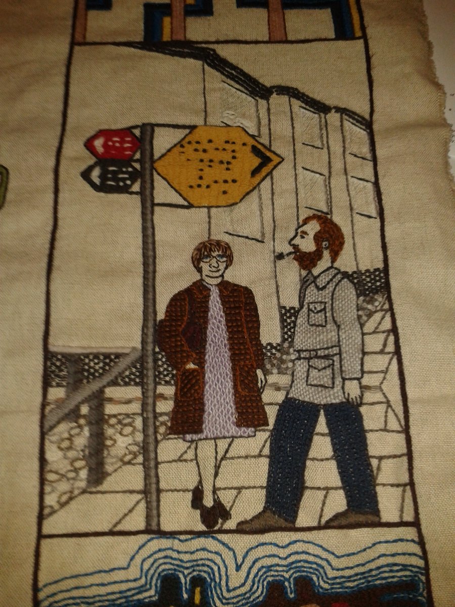 Detail from panel 140 of the Great Tapestry of Scotland (Cumbernauld New Town) showing my parents in the 1970s next to a stylised street sign with the words Sheila and Jim in Braille, and Mum and Dad in Morse code.  A memorial image of Snowflake the cat is in the window of the background houses.