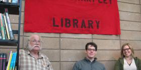 Kate Sharpley Library bookfair stall: three people beneath a red and back banner