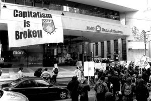 Giant "Capitalims is Broken" banner hangs from Bank of Montreal building while protestors gather below.