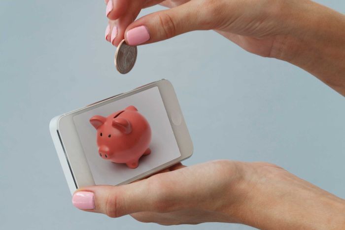 A woman holds a coin above a phone with an image of a piggy bank on it. 