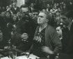 B&W Elderly woman in spectacles standing at a table with microphone, surrounded by seated people.