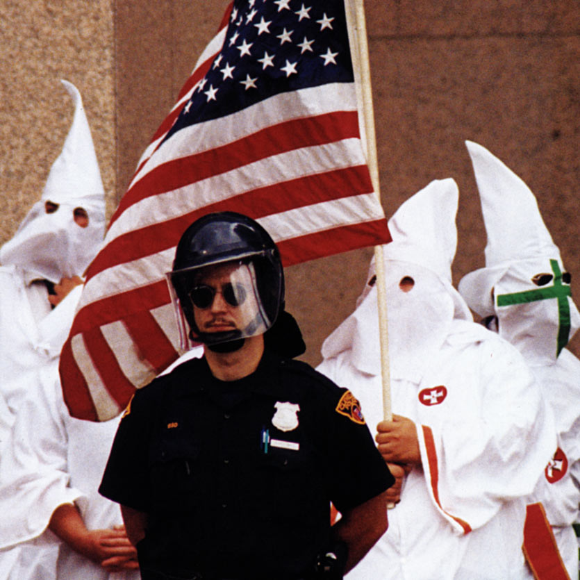a mustachioed cop in riot gear wearing aviator sunglasses stands guard in fron of severl Ku Klux Klan members in full robes, one of whom carries a American flag
