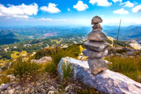 Stone cairns pyramid built on top of a mountain with a panoramic view of the Montenegrin valley of hills and forests under a blue sky with white clouds. Vidikovac, Lovcen National Park, Montenegro. Traveller Quiz February 8, 2019