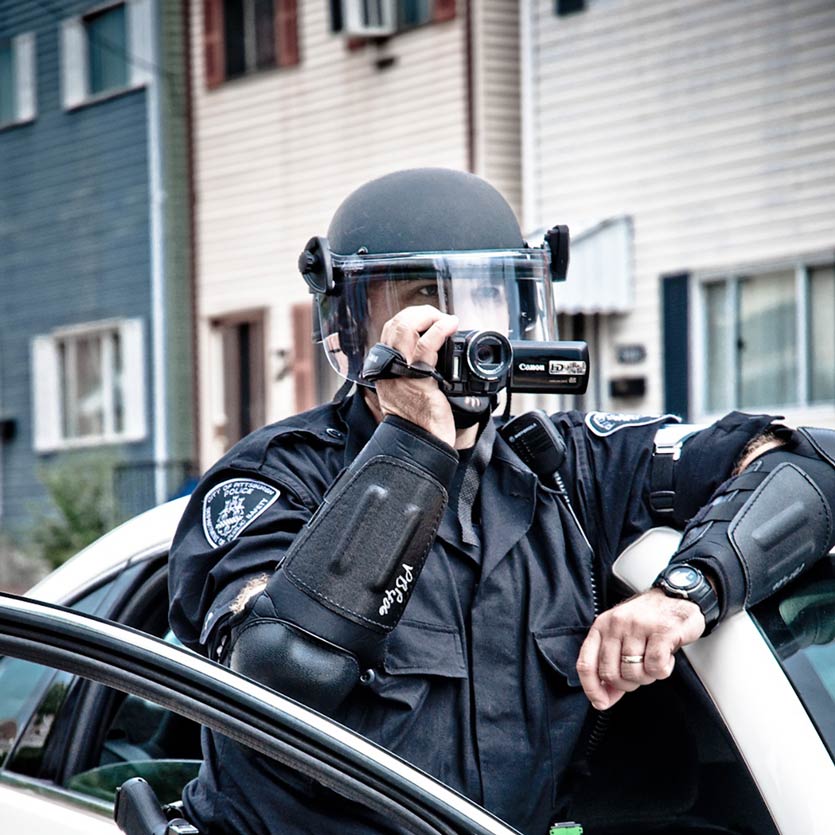 a cop dressed in fancy riot gear leans out of his open patrol car door and uses a camcorder to record protestors who are not pictured