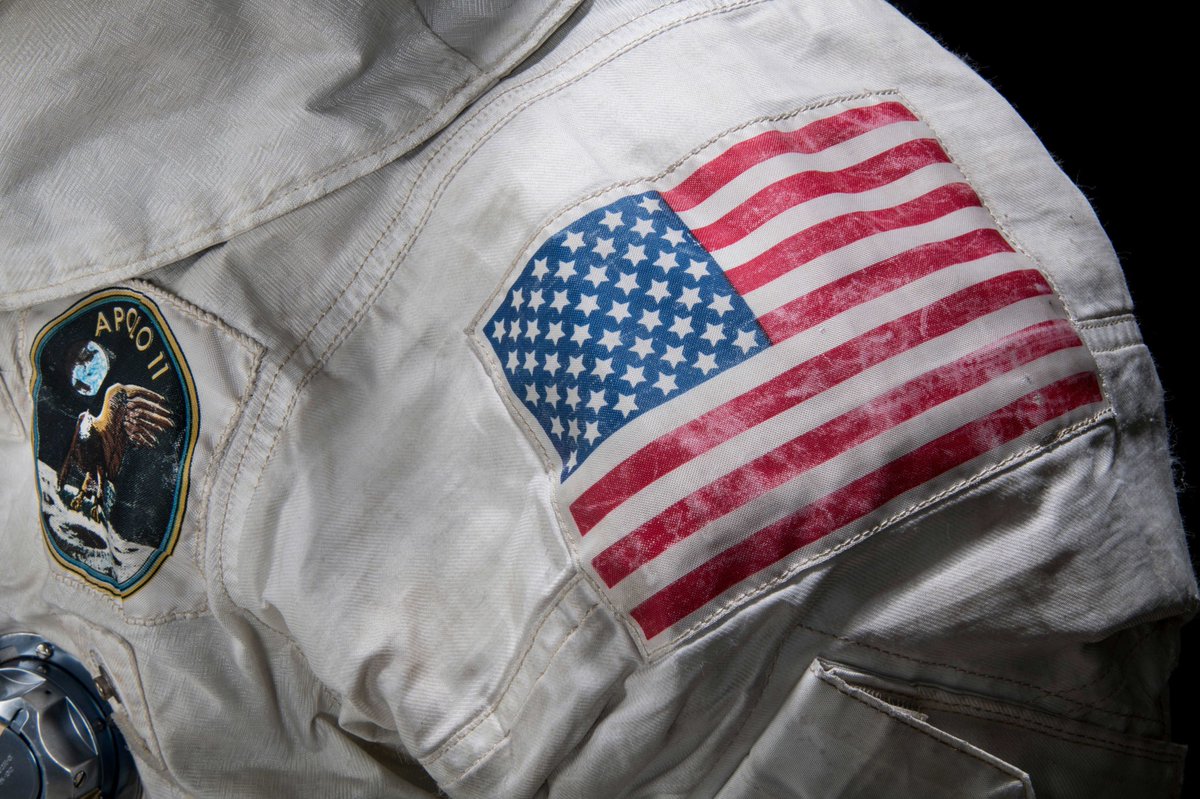 Close up photograph of the U.S. flag on the shoulder/arm portion of a white spacesuit. The flag appears slightly worn and aged. Also visible is an Apollo II emblem on the chest with an image of an eagle.