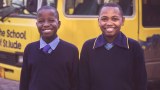 Two Tanzanian children stand in front of their school bus in uniform.