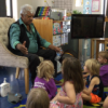 An Aboriginal elder sits in a chair speaking to a room for preschool children.