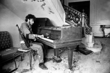 A black and white photograph of a man with a rifle slung over his shoulder playing the piano