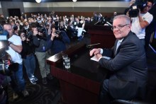 Morrison sits behind a lectern in front of a wall of people with cameras.