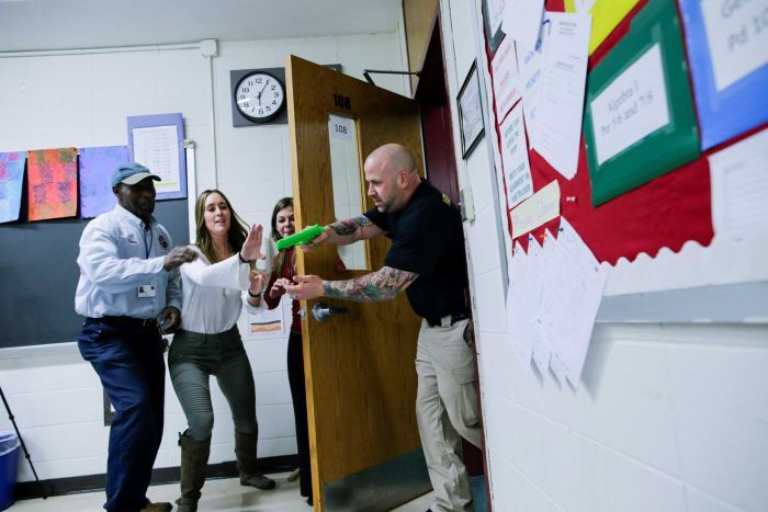 A man holding a green plastic gun bursting into a classroom while a teach tries to stop him 