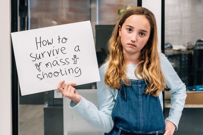 A girl in overalls holding a sign that reads "How to survive a mass shooting" 