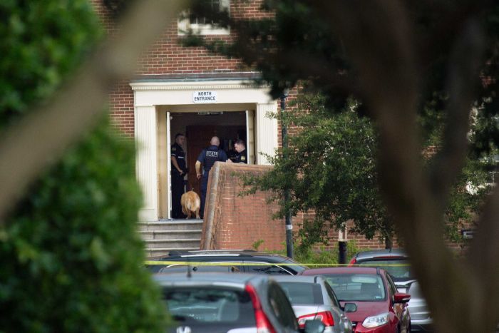 Three police officers and a police dog stand at the entrance of a brown brick building.