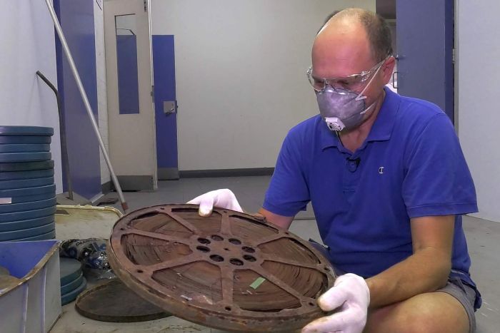 Paul Lyons sits holding a huge spool of film while wearing white gloves and a protective breathing mask. 