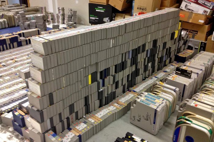 Hundreds of television video tapes in grey boxes stacked in an empty room. 