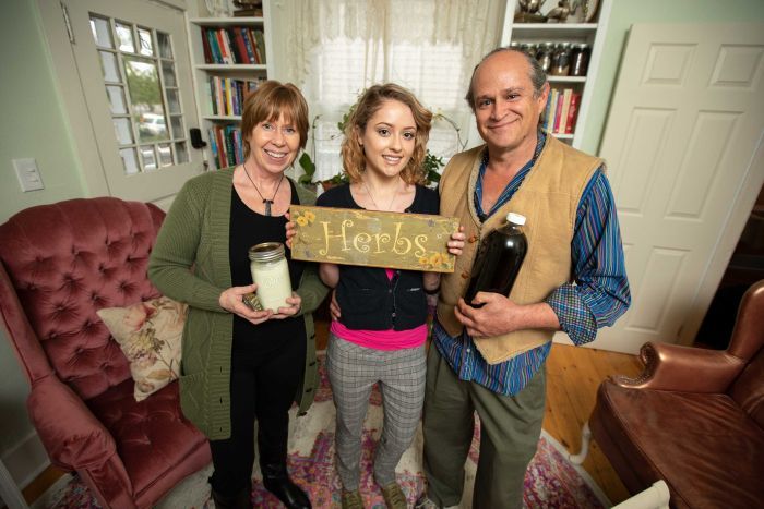 A young woman holds a sign reading herbs, while an older woman holds a candle and a man holds a bottle of dark liquid