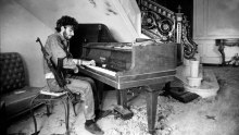 A black and white photograph of a man with a rifle slung over his shoulder playing the piano