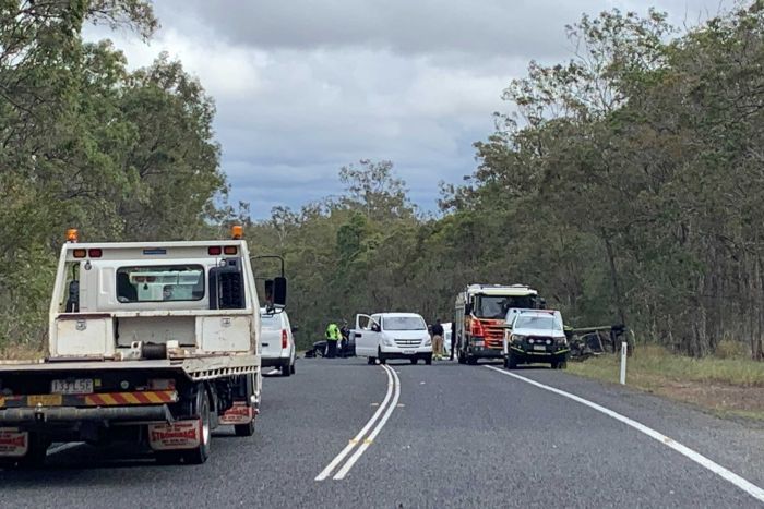 The wreckage of three crashed cars sits surrounded by police and other cars at the scene of a crash on a tree lined highway.