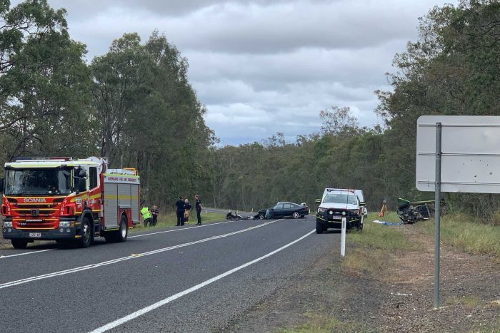 The wreckage of three crashed cars sits surrounded by police and other cars at the scene of a crash on a tree lined highway.