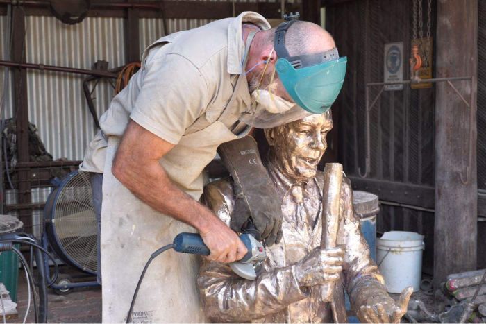 A man wearing a protective mask over his mouth and shield over his eyes buffs a bronze statue of a man with a power tool.