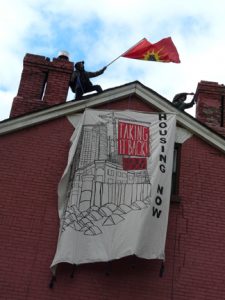 Giant Taking it Back: Housing Now banner hanging from roof of empty building at 230 Sherbourne. Above stand two Indigenous men, one waves the Unity flag. 