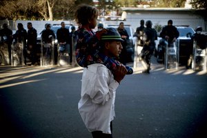 A migrant carries a child past Mexican police who stand guard outside the Benito Juarez Sports Center which is serving as a shelter for migrants in Tijuana, Mexico, Monday, Nov. 26, 2018.