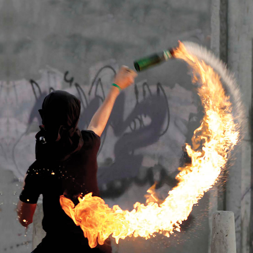 a protestor with his face concealed in the act of throwing a lit molotov cocktail during a protest, in front of a huge concrete wall