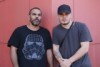 Two Indigenous men standing next to each other with a red wall in the background.