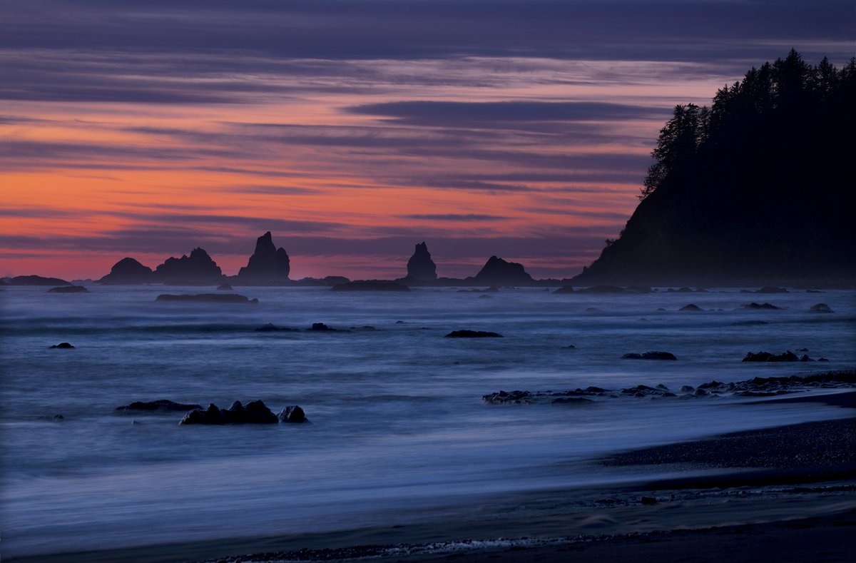 Ocean waves wash up on a beach with rocks rising out of the water under a colorful sunset sky.