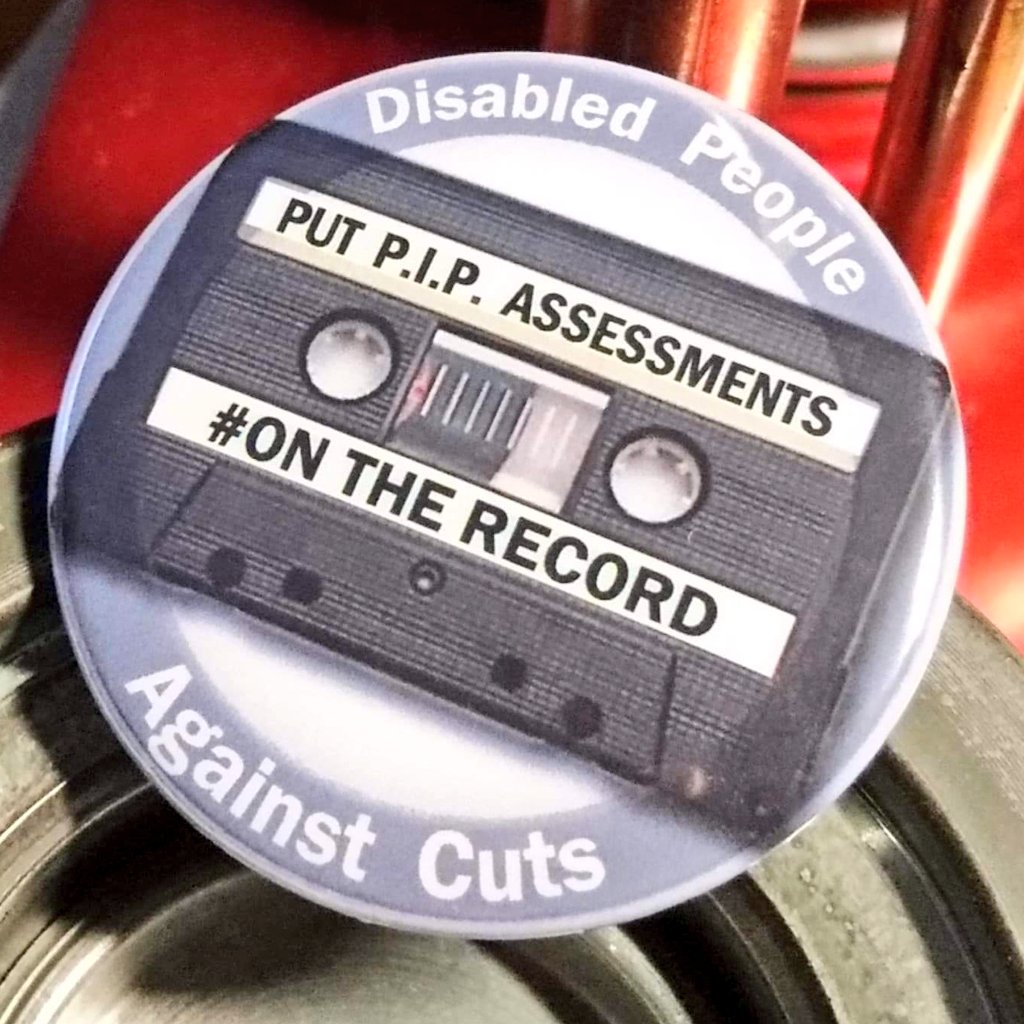 an image of a badge which is circular around the edge it says disabled people against cuts across the middle is the image of a cassette tape on the cassette tape it says put pip assessments on the record