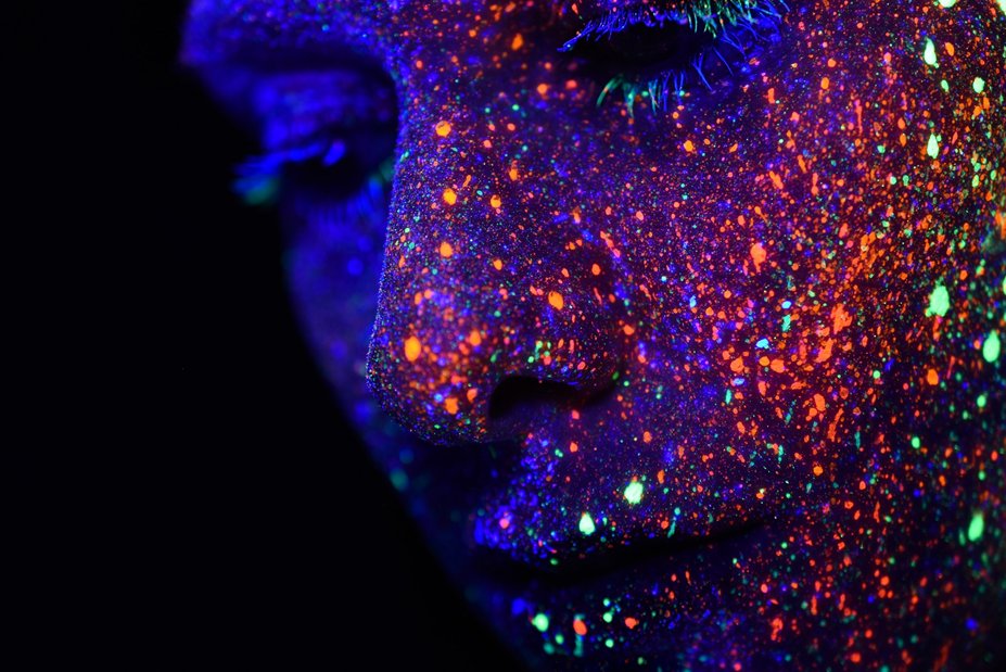 Close-up photo of human face, eyes closed, in semi-profile against dark background. Face is speckled with multicolored fluorescent paint. Image credit: H. Heyerlein via Unsplash.