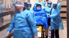 A man on a gurney wears a mask as doctors in larger masks push him through a hospital corridor.