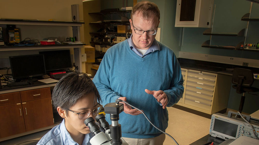 A man stands in a lab behind another researcher in a lab coat who is using a microscope