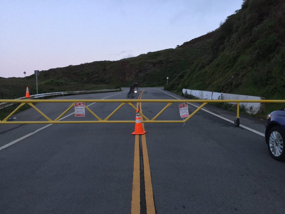 Temporary yellow metal barricade blocking the full width of Twin Peaks Boulevard with an orange cone in front of it, and NO STOPPING TOW AWAY signs attached to it in a couple of places, behind it a bicyclist swerving towards the right edge of the gate, and the hill up to Twin Peaks itself.